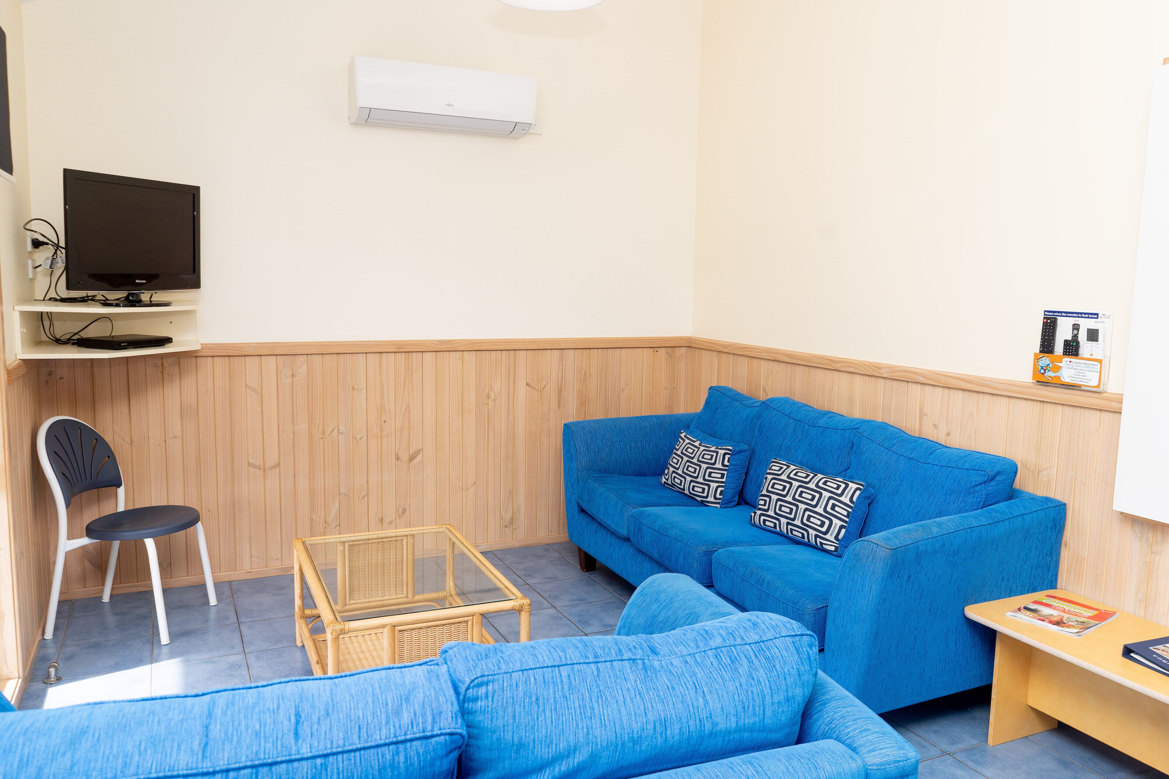 Quarto com sofá azul, uma cadeira, uma mesinha, televisão e aparelho de ar condicionado.