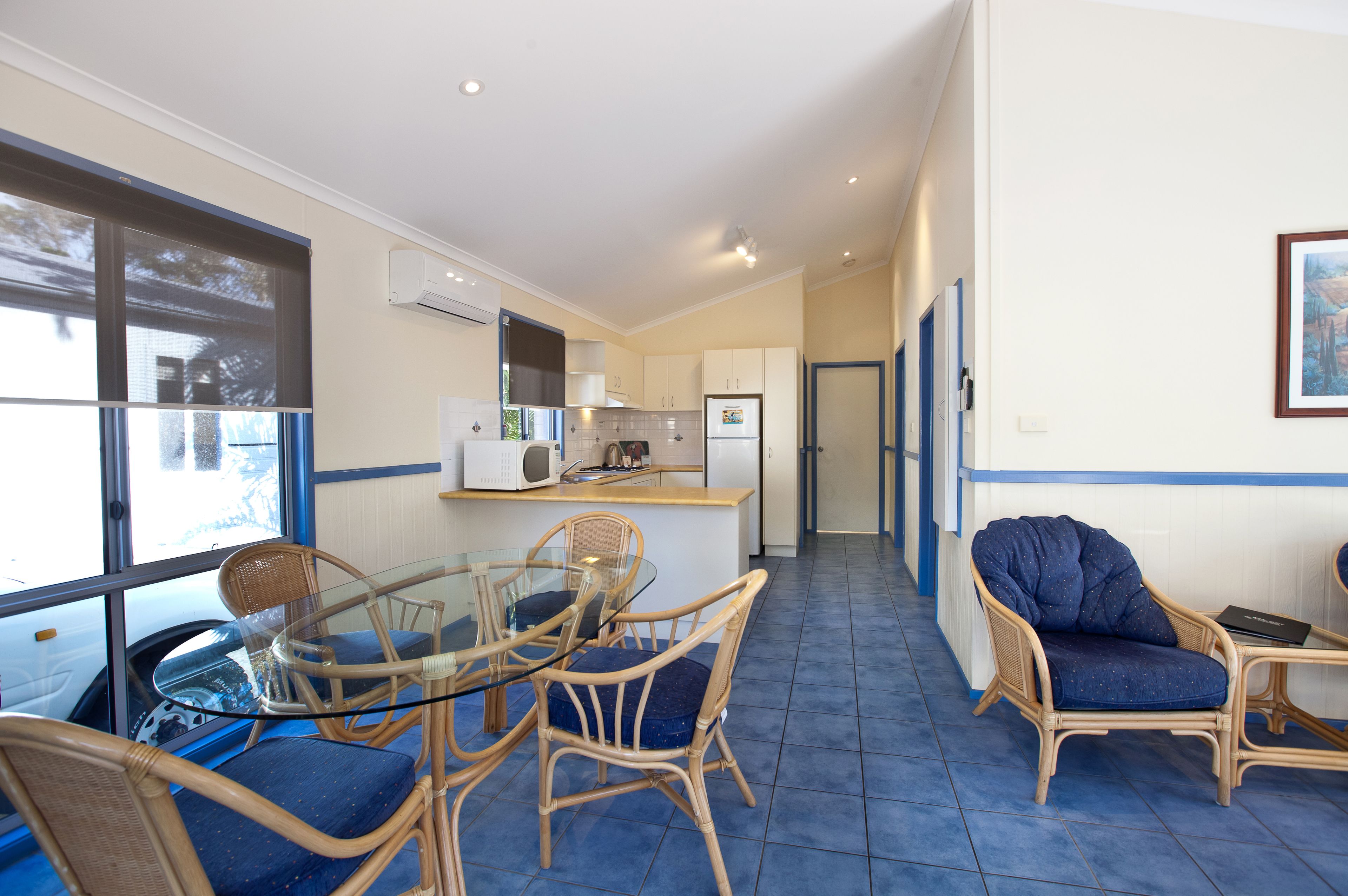 A compact kitchen with white cabinets, a microwave, and a refrigerator. There is a dining area with wicker chairs and a glass table.