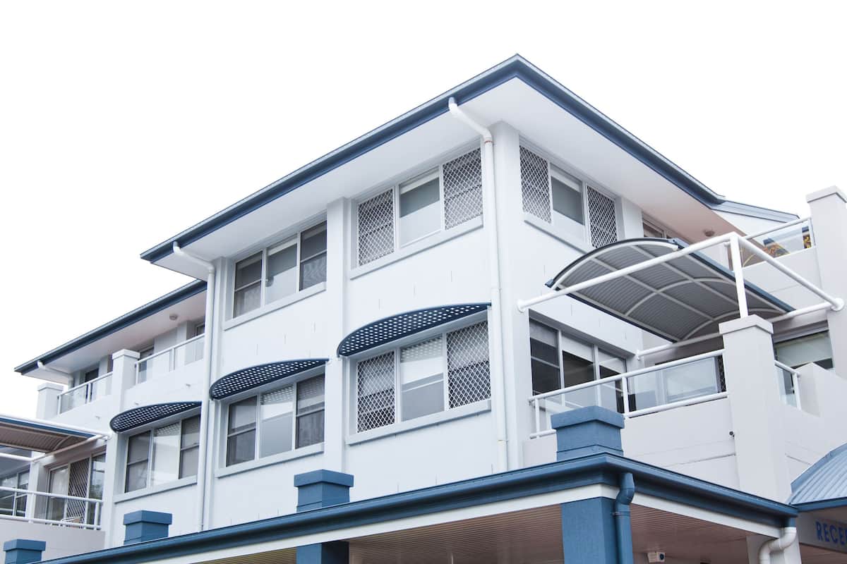 A multi-story building with a white exterior and blue trim. It features large windows with mesh screens, balconies with metal railings, and a covered outdoor area.