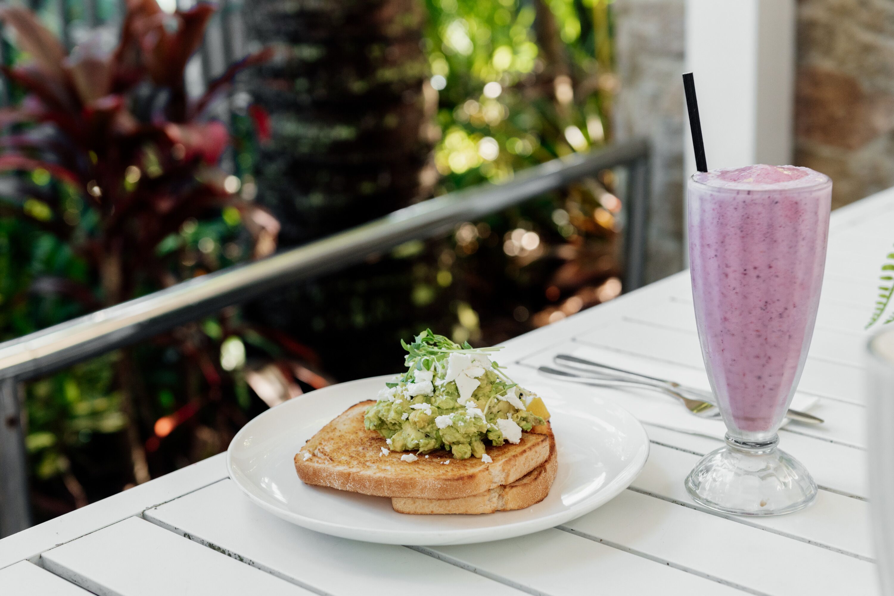 A plate with toast topped with avocado and greens, and a glass of smoothie on a white table.