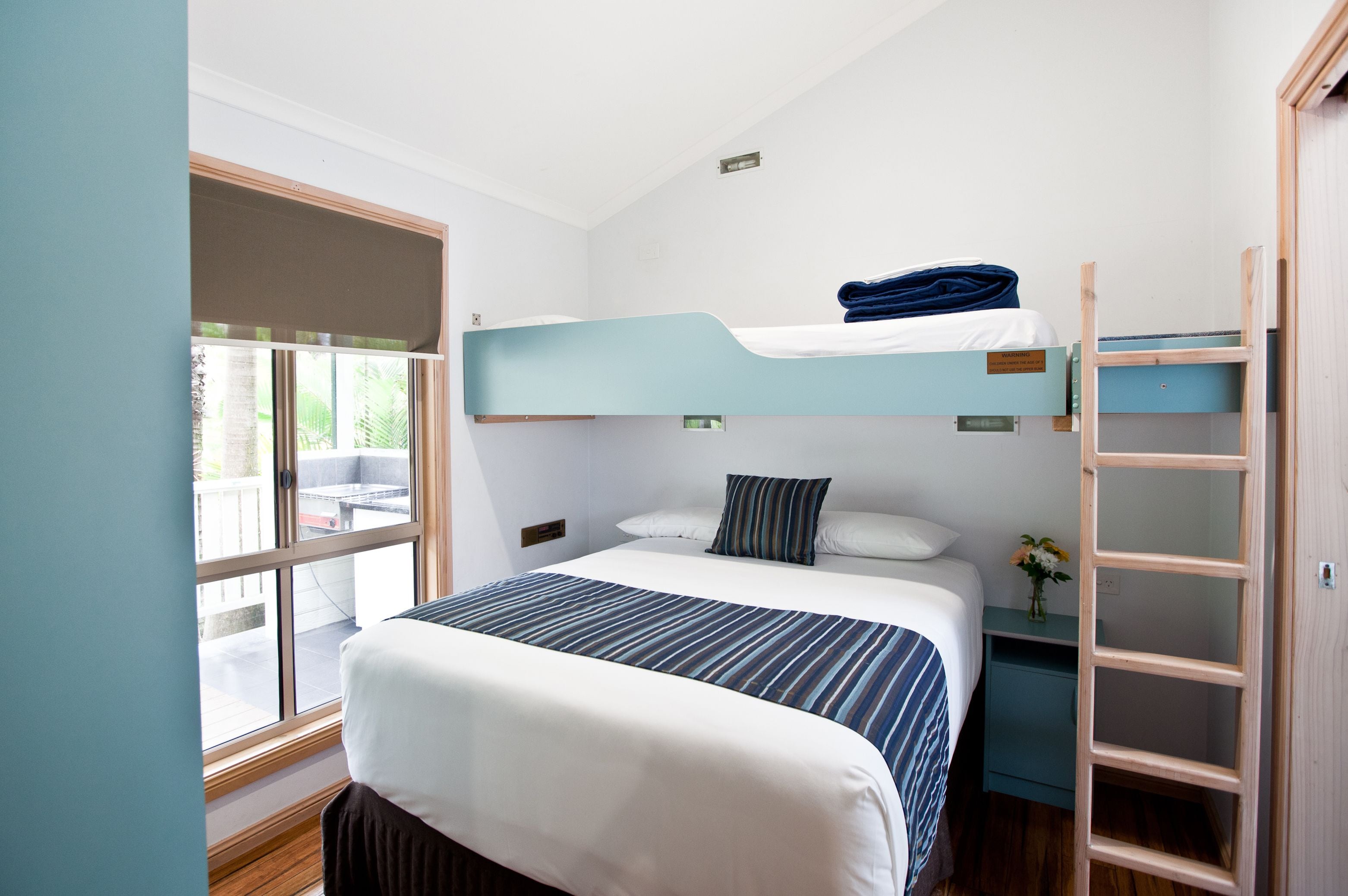 A bunk bed room with a window, a bedside table with a vase, and a wooden ladder.