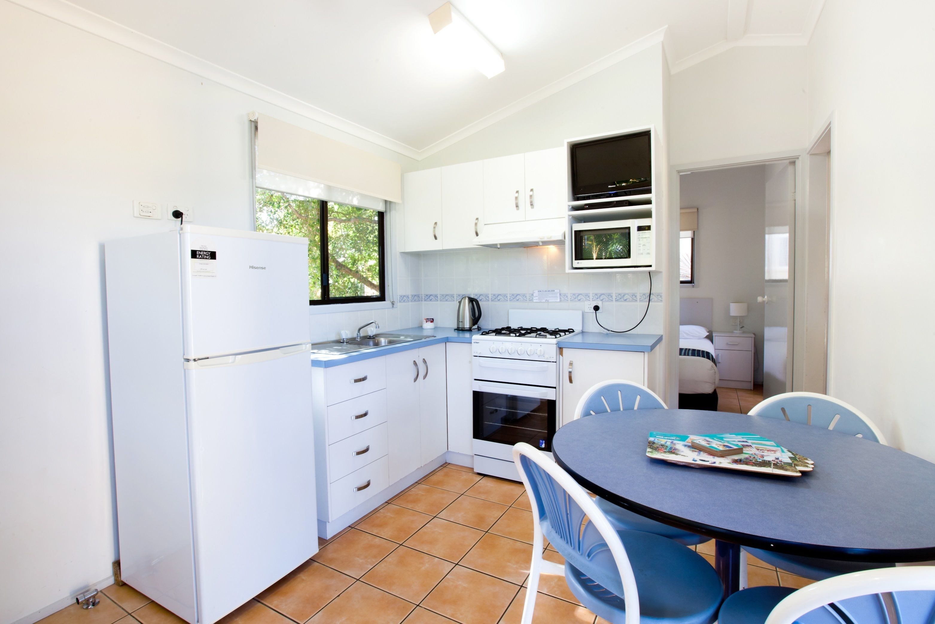 Cocina compacta con armarios blancos, un refrigerador, estufa y microondas. Cuenta con una mesa de comedor con sillas y una ventana que proporciona luz natural.