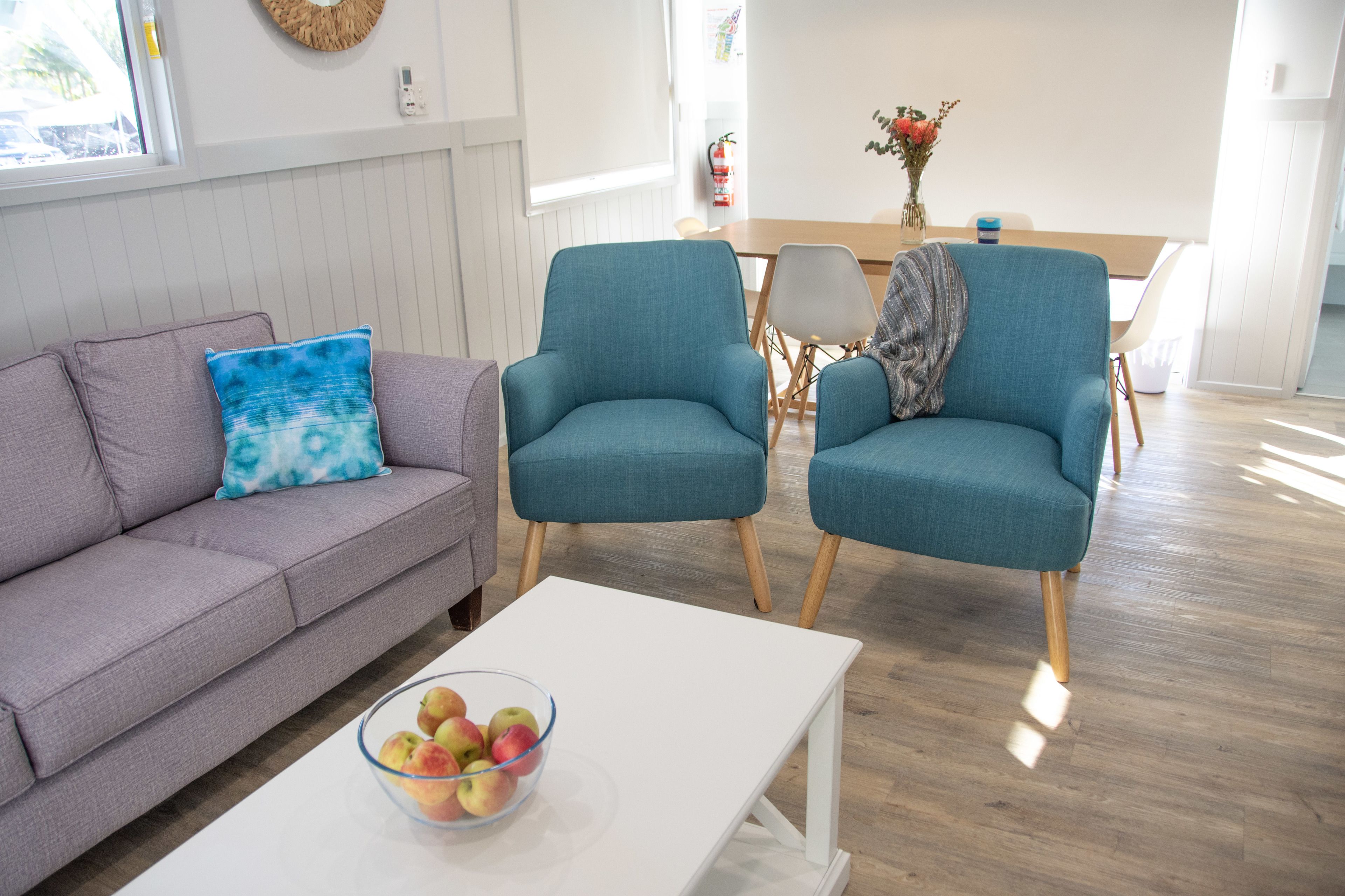 A modern living room with a grey sofa, teal armchairs, a white coffee table with a bowl of apples, and a wooden dining table with chairs.
