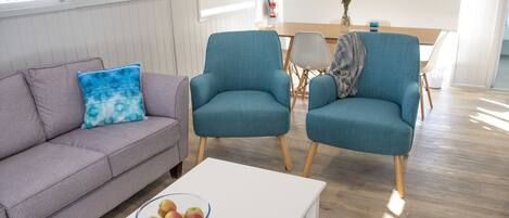 A modern living room with a grey sofa, teal armchairs, a white coffee table with a bowl of apples, and a wooden dining table with chairs.