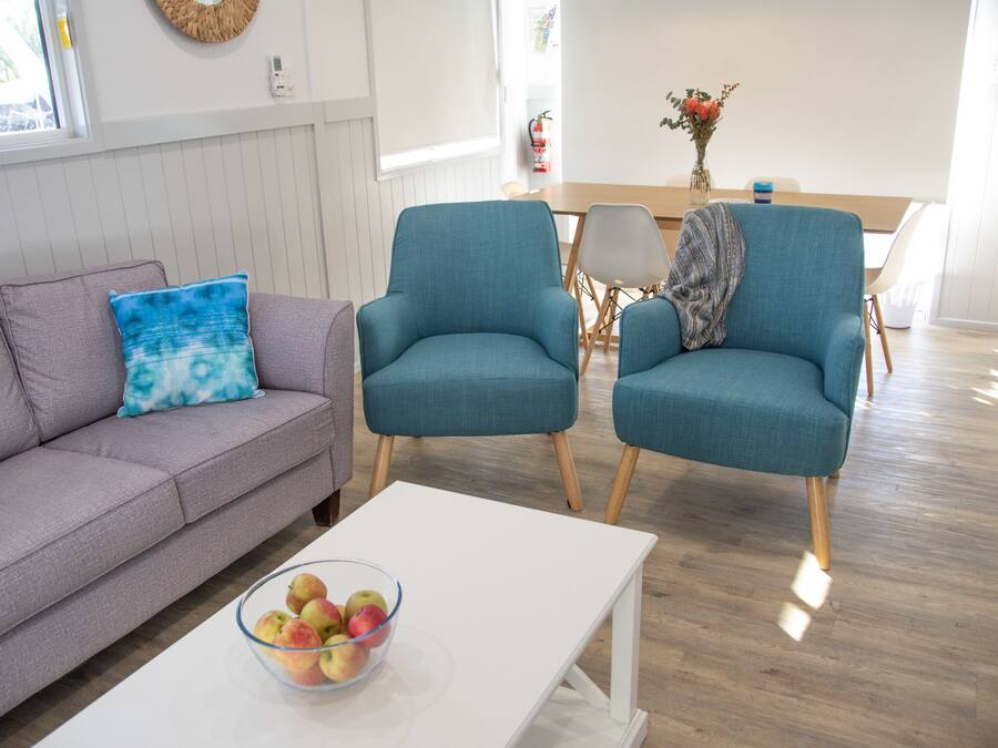 A modern living room with a grey sofa, teal armchairs, a white coffee table with a bowl of apples, and a wooden dining table with chairs.