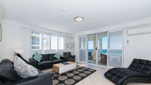 A spacious living room with a black leather sofa, a wooden coffee table, and a black leather armchair. There is a large window with blinds, a sliding glass door leading to a balcony, and a white ceiling with a recessed light fixture. - La Grande Apartments Broadbeach (Broadbeach)