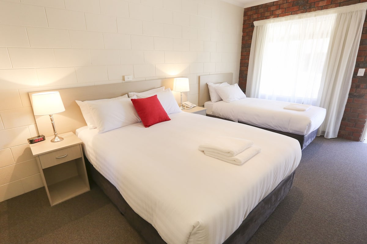 Ett hotellrum med två sängar, vita sängkläder, en röd kudde, sänglampor, ett nattduksbord med en telefon och en vägg av tegelsten.