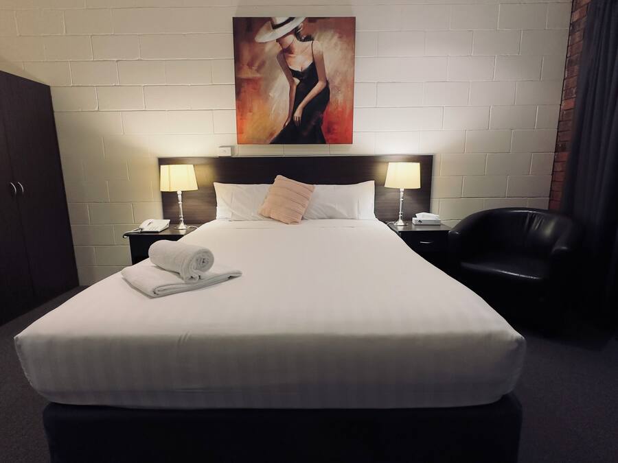 A hotel room with a neatly made bed, a headboard painting, a desk with a lamp, a chair, and a wardrobe.