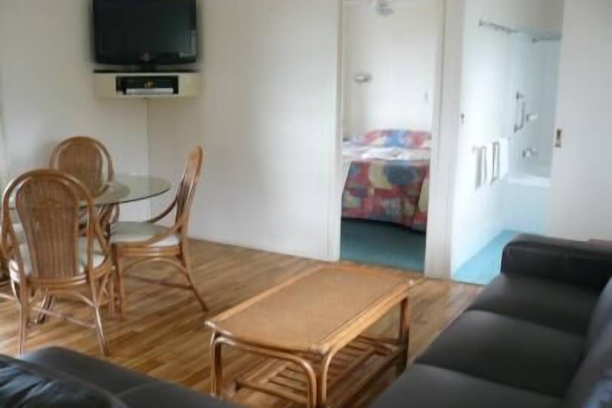 Un espacio habitable compacto con un sofá, una mesita y dos sillas. Un televisor está montado en la pared, y se ve una cama con una manta colorida a través de una puerta abierta.