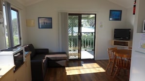 A compact living space with a kitchenette, a sofa, a dining table with chairs, a television, and a sliding glass door leading to an outdoor area. - Diamond Beach Holiday Park (Diamond Beach)