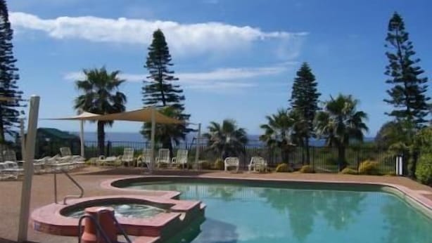 A pool with lounge chairs and umbrellas, surrounded by palm trees and tall conifers. - Diamond Beach Holiday Park (Diamond Beach)