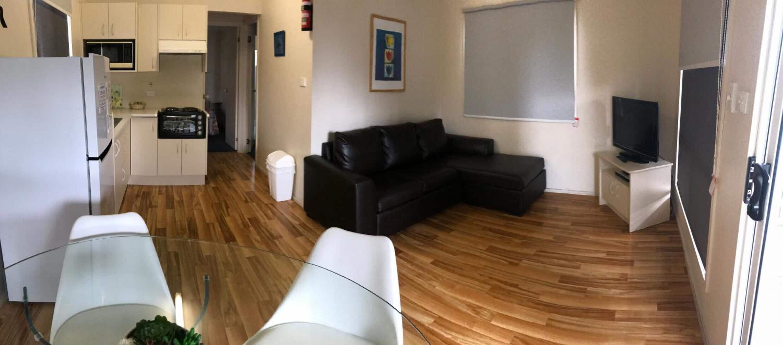 A compact living space with a kitchenette, a black leather sofa, a glass dining table, and a television.