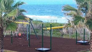 A playground with swings and a netted climbing area, surrounded by palm trees and overlooking the ocean. - Diamond Beach Holiday Park (Diamond Beach)