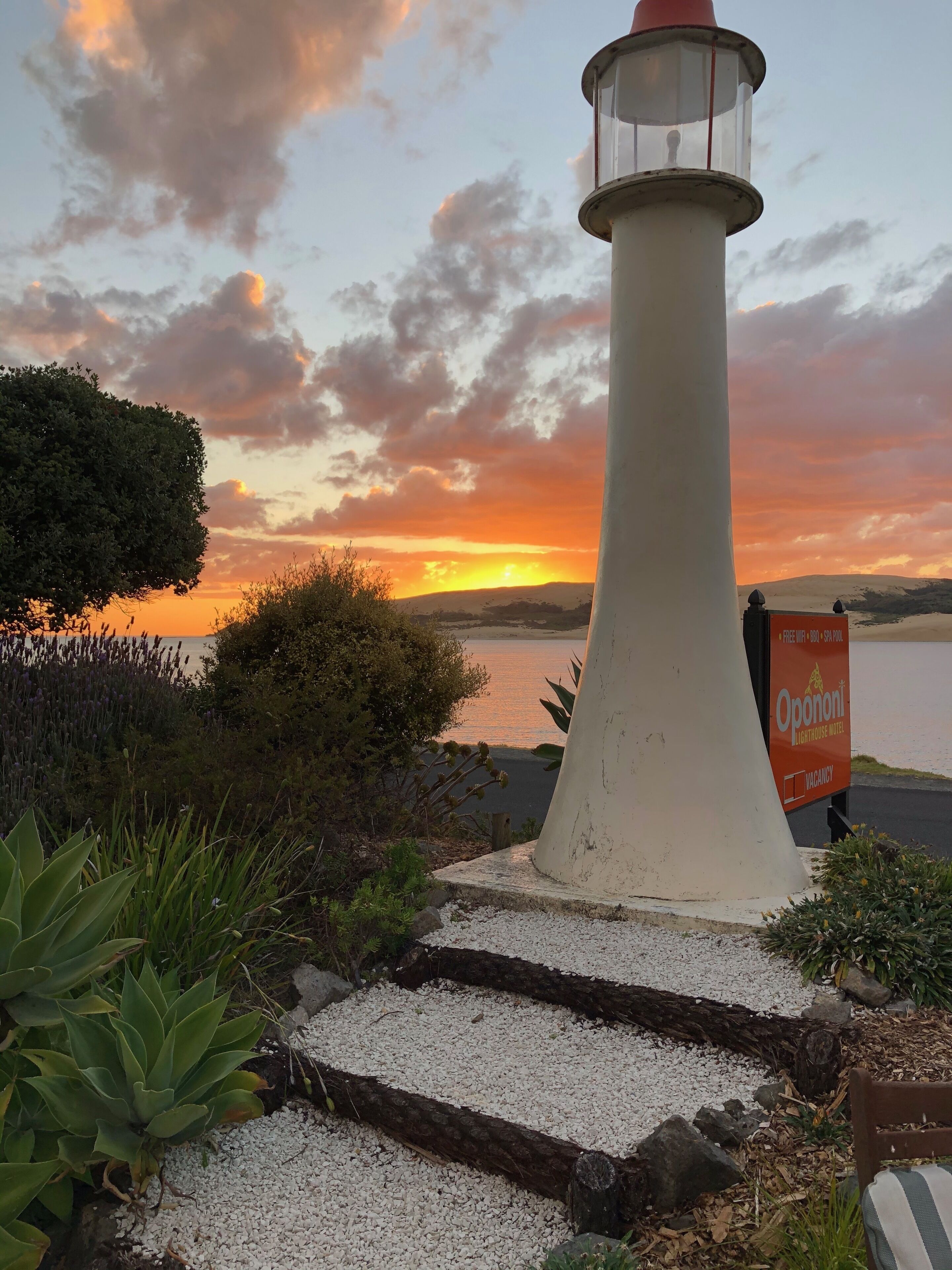 A lighthouse with a sign indicating vacancy, surrounded by vegetation and a sunset view.