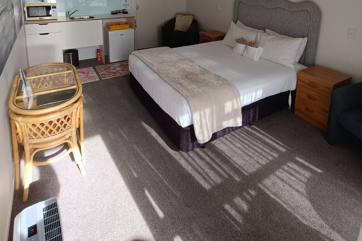 Une chambre d’hôtel comprenant un lit, des tables de chevet, un petit bureau avec une chaise, un téléviseur fixé au mur et un ventilateur de plafond.