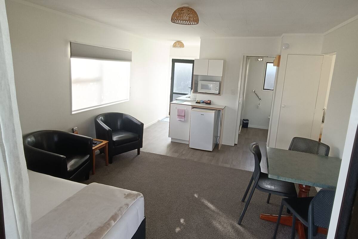 Une chambre d’hôtel compacte comprenant un lit, une petite kitchenette, un coin repas avec une table et des chaises, et un coin salon avec deux fauteuils.