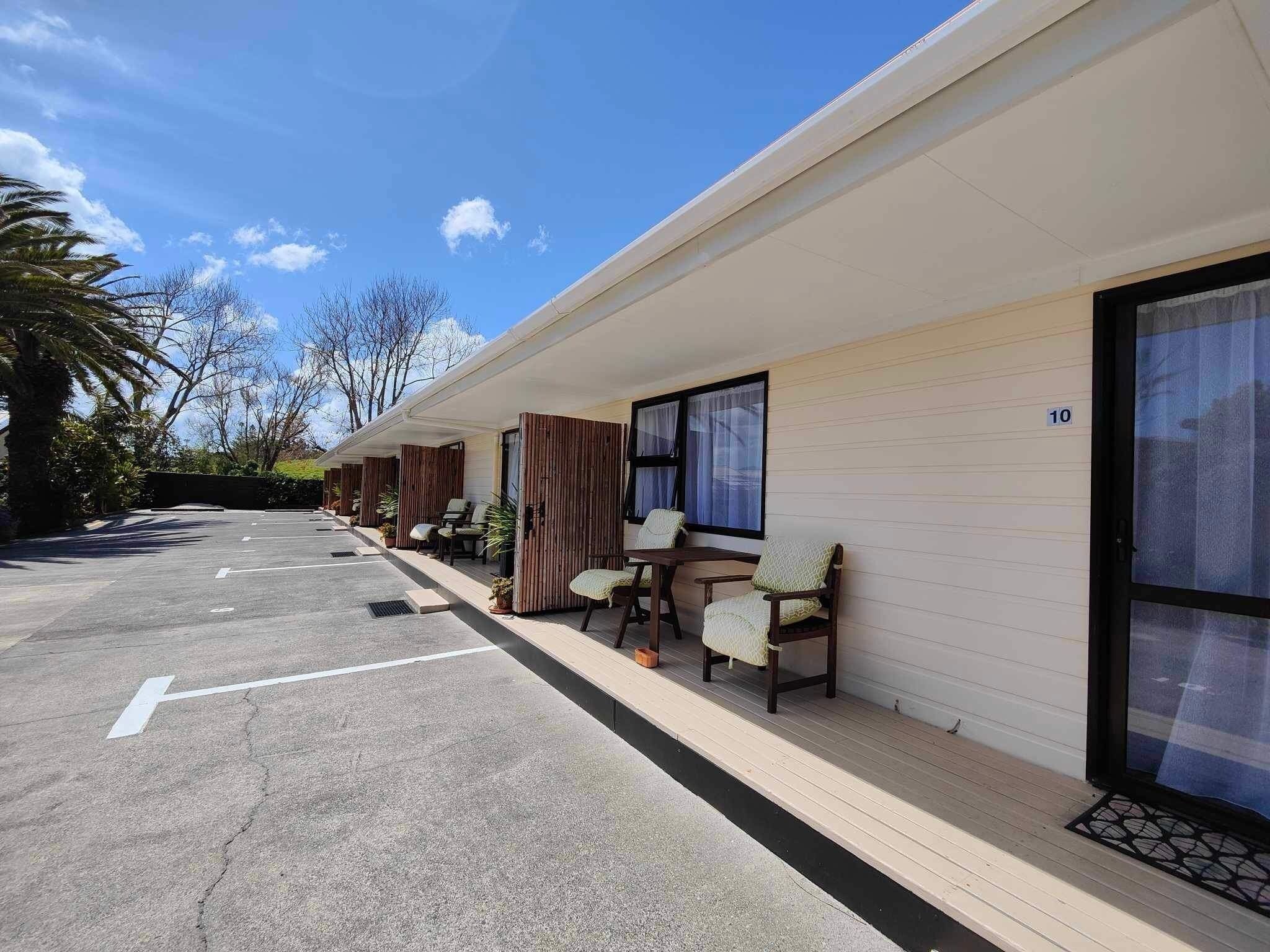 A row of motel rooms with individual windows, a covered outdoor area with chairs, and a parking lot.