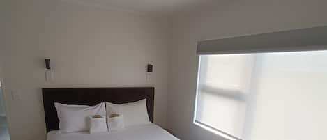 Una cama bien hecha con sábanas blancas en una habitación con un ventanal grande, cabecera oscura y una lámpara colgante.