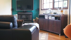 A living room with a leather sofa, a flat-screen TV, a fireplace, a cabinet with books, and a window with natural light. - Yallambee (Bundanoon)
