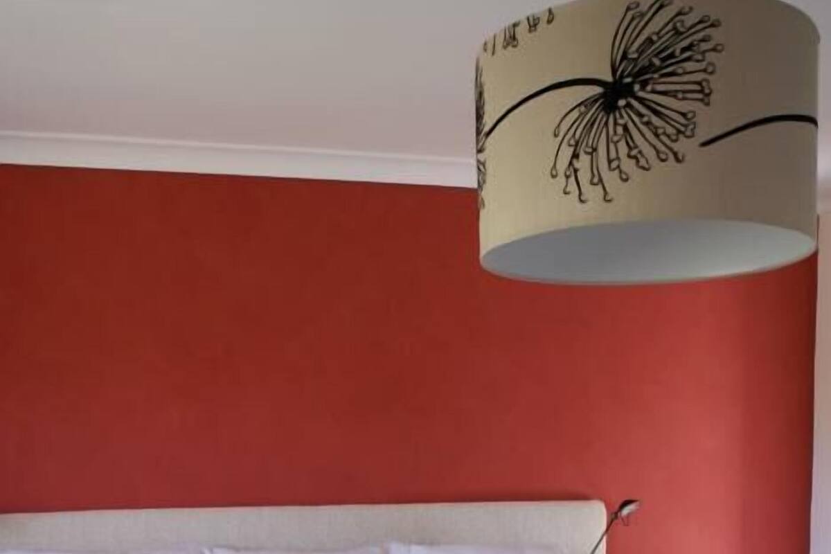 Cama bem arrumada com almofadas brancas e vermelhas, uma luminária em uma mesa de cabeceira e uma parede vermelha.