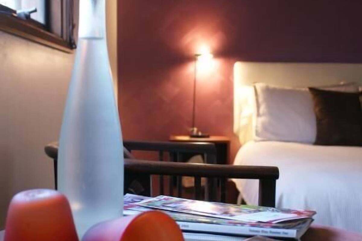 Quarto de hotel com cama, mesa de cabeceira, abajur, uma garrafa e algumas revistas sobre uma mesa.