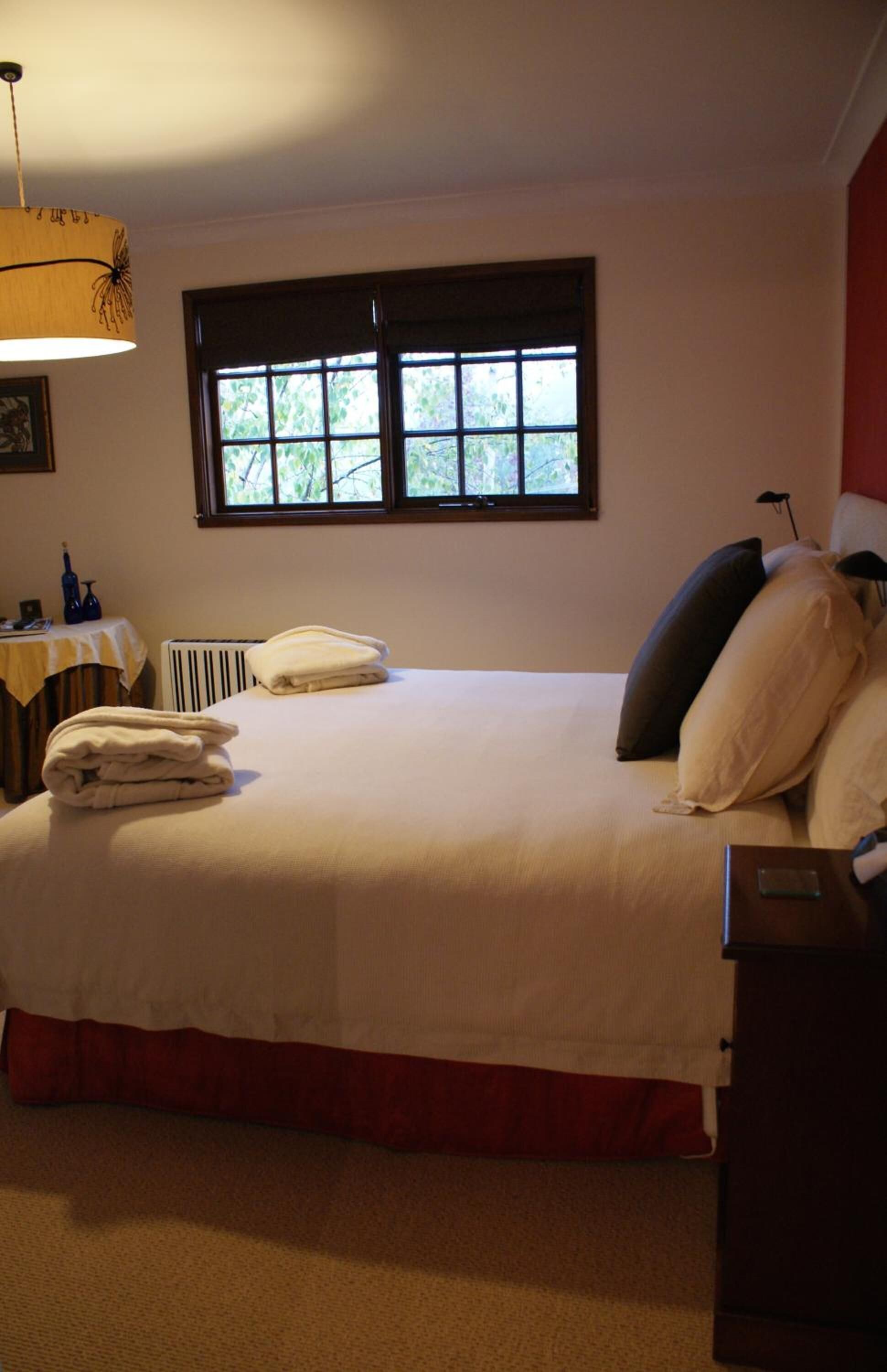 A neatly made bed with folded towels, a window with a view of trees, a table with a lamp, and a radiator.