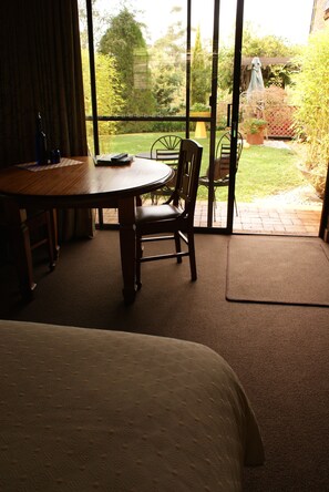 A room with a bed, a wooden table, and a chair. There is a window with a view of a garden.