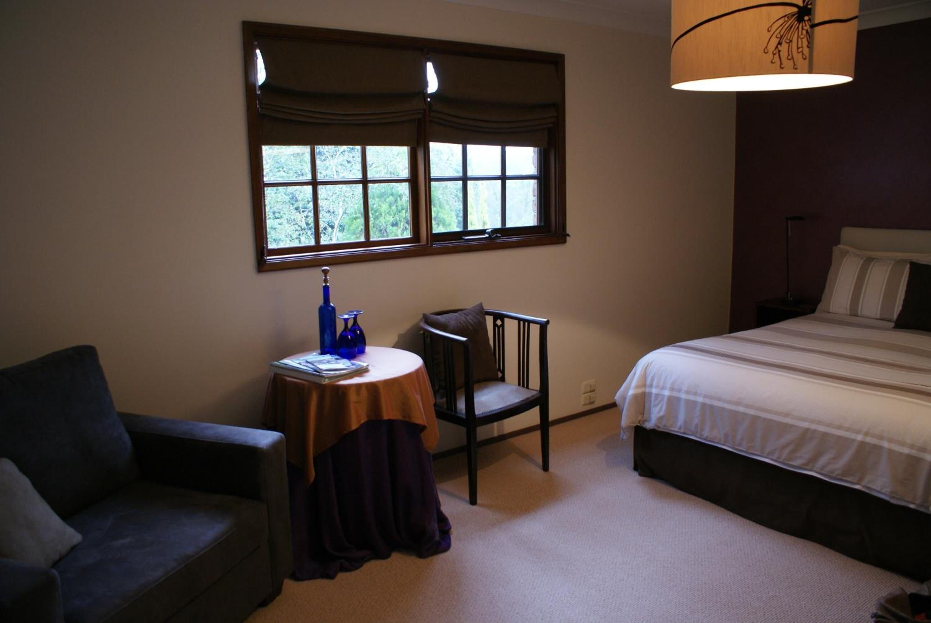 A bedroom with a bed, a chair, a small table with a lamp, and a window with curtains.