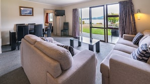 A living room with a sofa, a coffee table, and a TV. There is a large window with curtains and a view of a lake. - Lakeside Motel & Apartments (Te Anau)