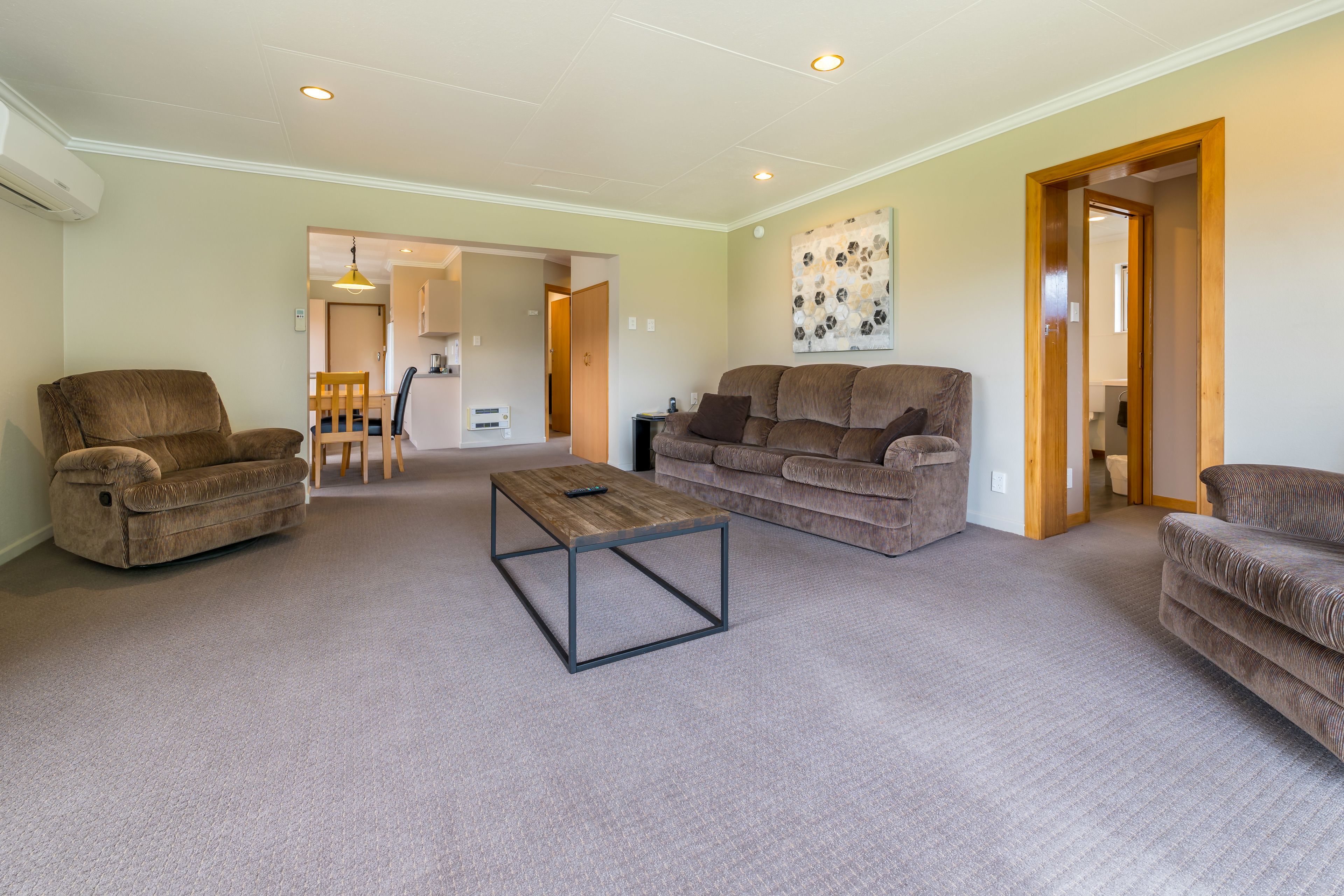 A spacious living room with a brown sofa, a wooden coffee table, and a brown armchair. There is a dining area in the background with a wooden table and chairs.