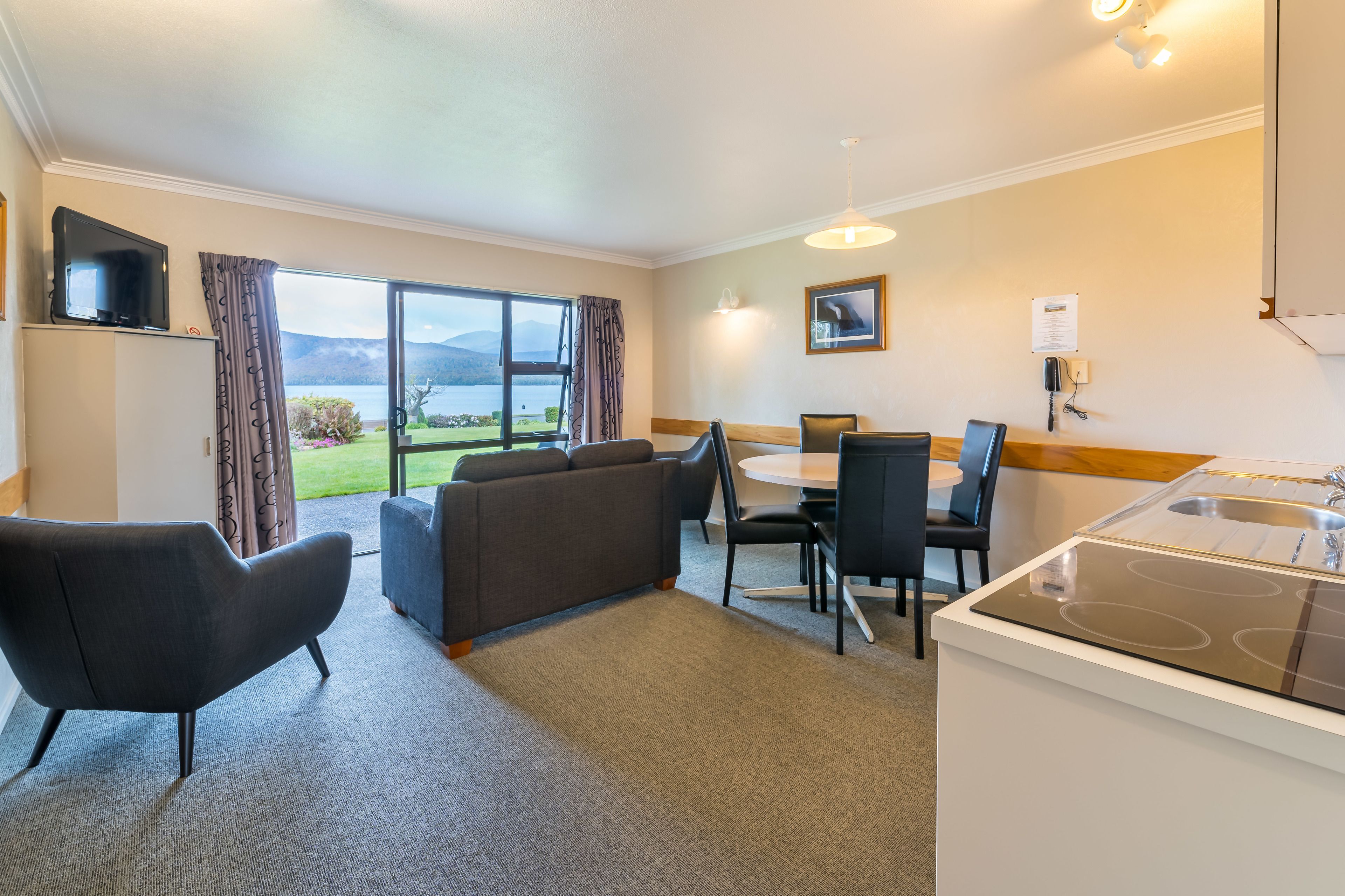 A modern hotel room with a sofa, dining table, chairs, a kitchen area with a stove, and a large window offering a view of the outdoors.