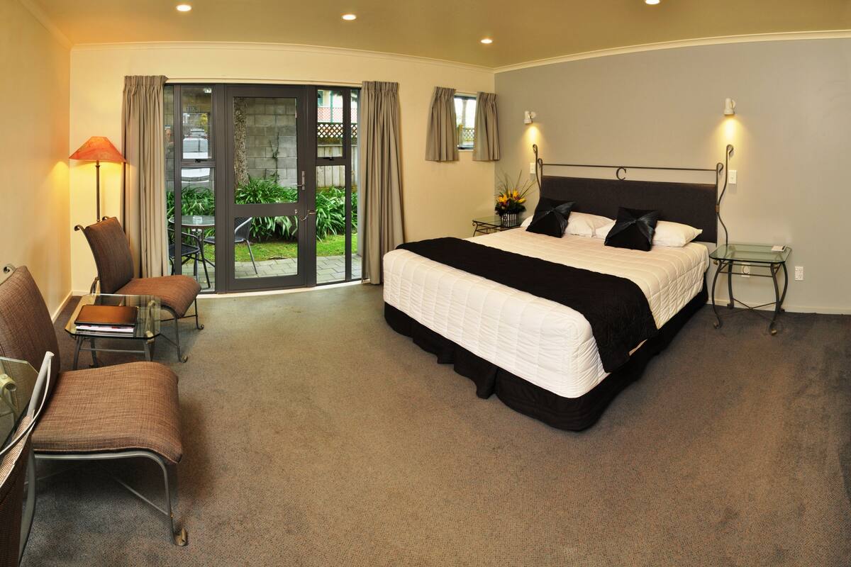 A hotel room with a large bed, a desk, a chair, a lamp, and a view of the outdoors through a sliding door.