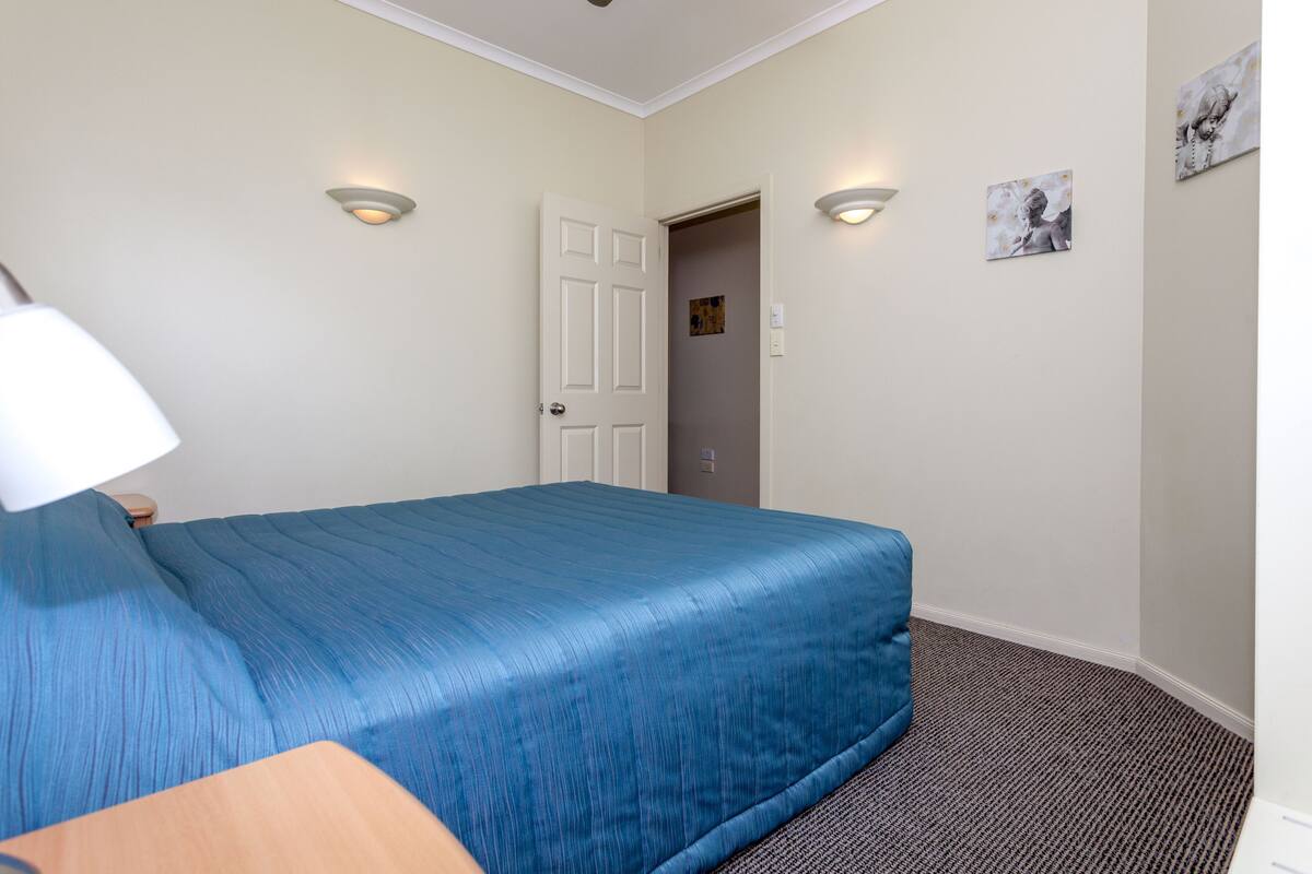 Una habitación de hotel con una cama, una mesita de noche, una lámpara y una puerta que da a otra habitación.