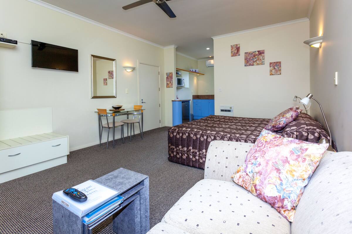 Habitación de hotel con cama, sofá, escritorio con control remoto, espejo y una kitchenette al fondo.