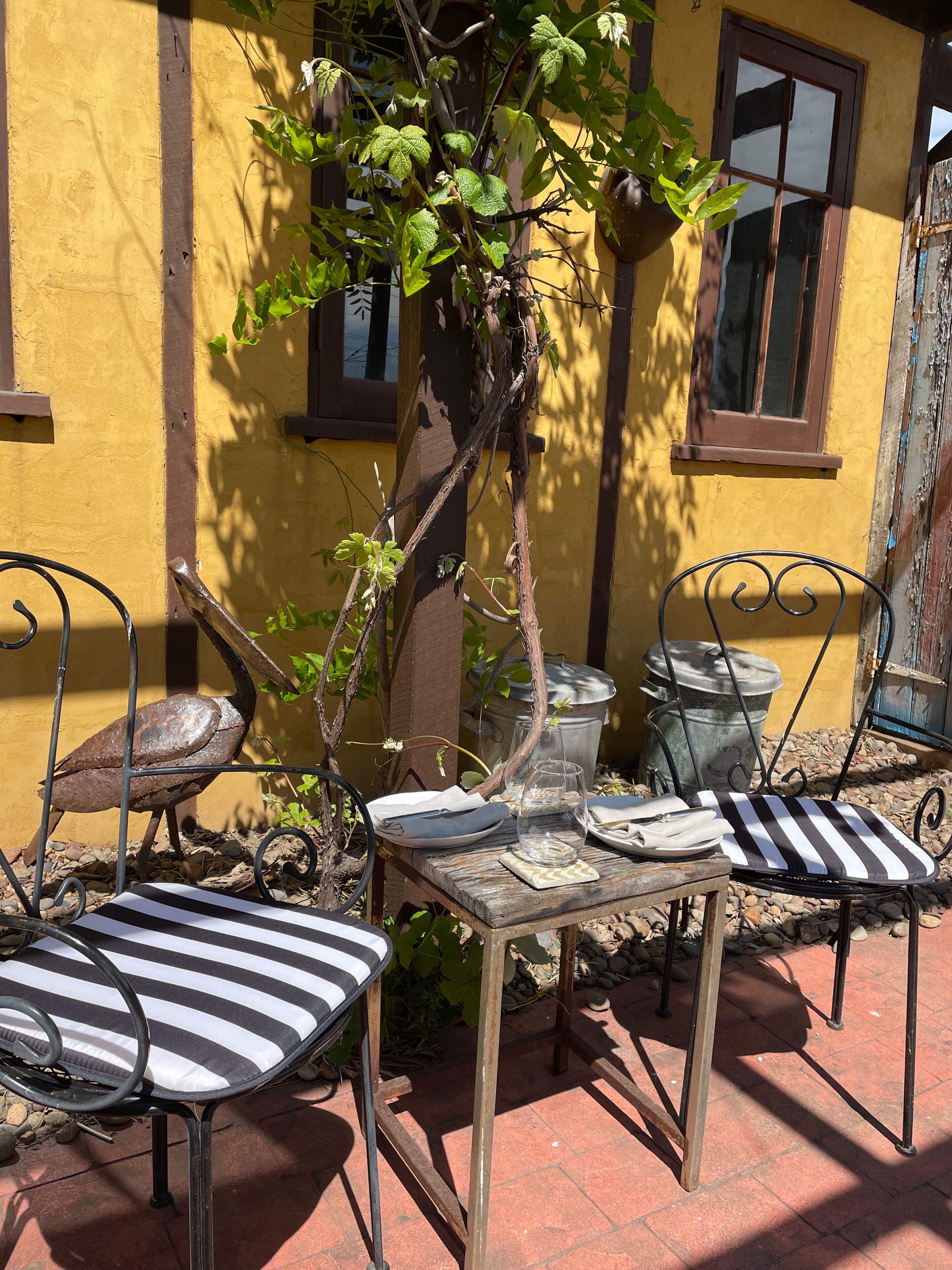 A patio with metal chairs and a table, a small tree, and a yellow wall with windows.