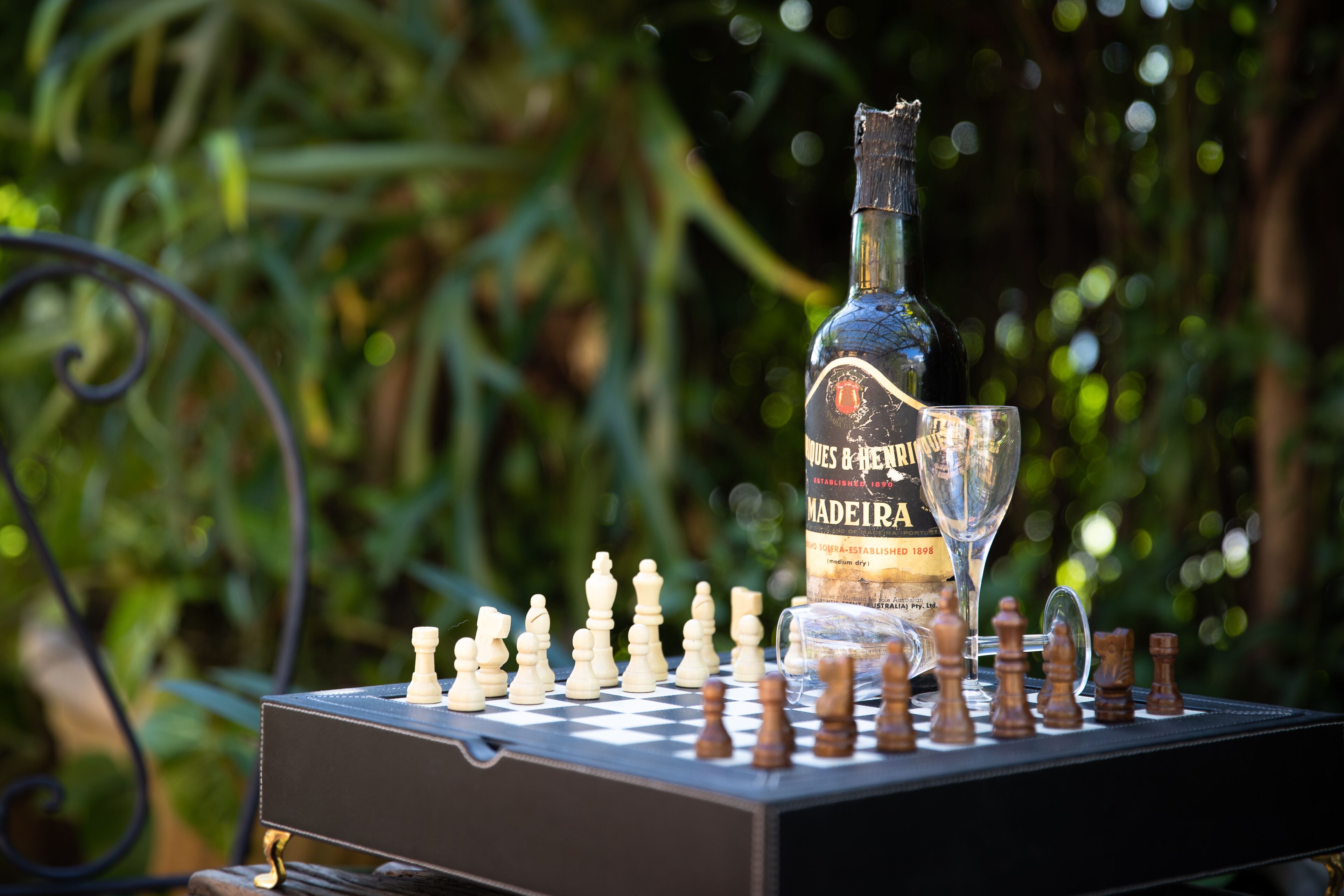 A chessboard with pieces, a bottle, and a glass on a table outdoors.
