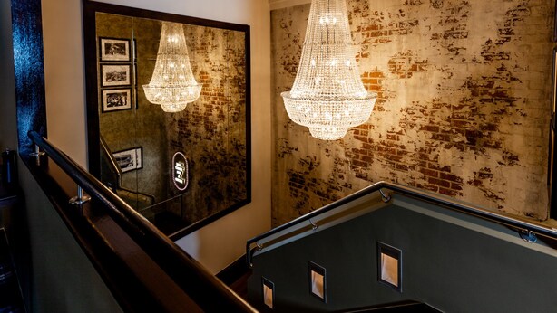 A staircase with a dark wooden railing, a wall with a distressed brick pattern, and two chandeliers. - Hotel Coronation (Sydney)