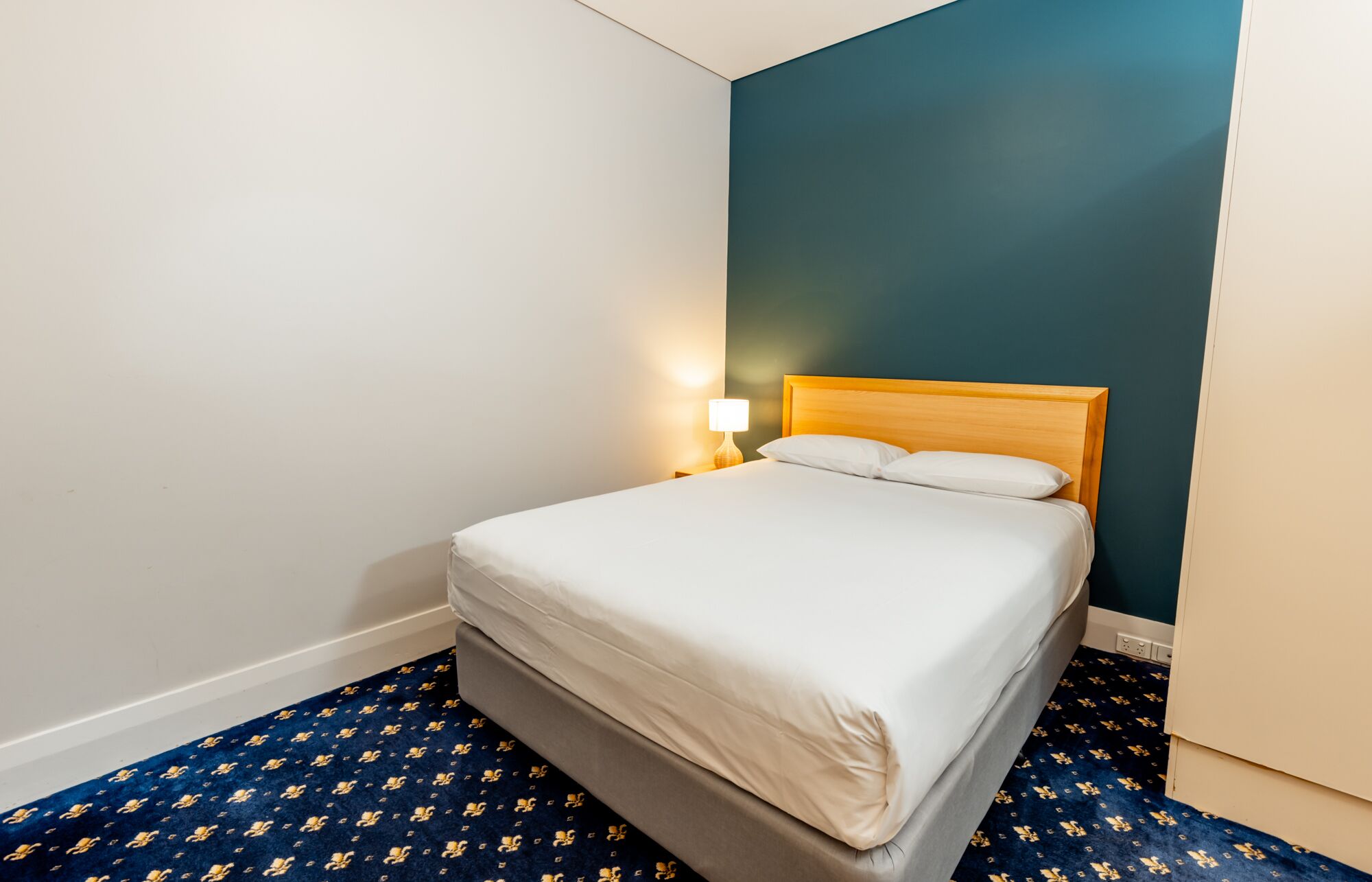 Una cama individual con cabecera de madera en una habitación con pared azul oscuro y alfombra estampada.