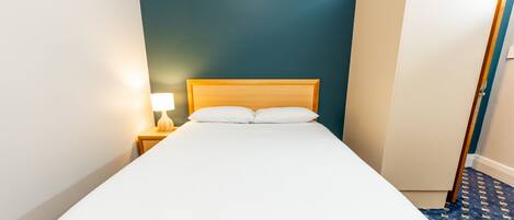 Una cama bien hecha con sábanas blancas en una habitación con paredes color turquesa, un cabecero de madera y una mesita de noche con una lámpara.