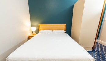 Una cama bien hecha con sábanas blancas en una habitación con paredes color turquesa, un cabecero de madera y una mesita de noche con una lámpara.