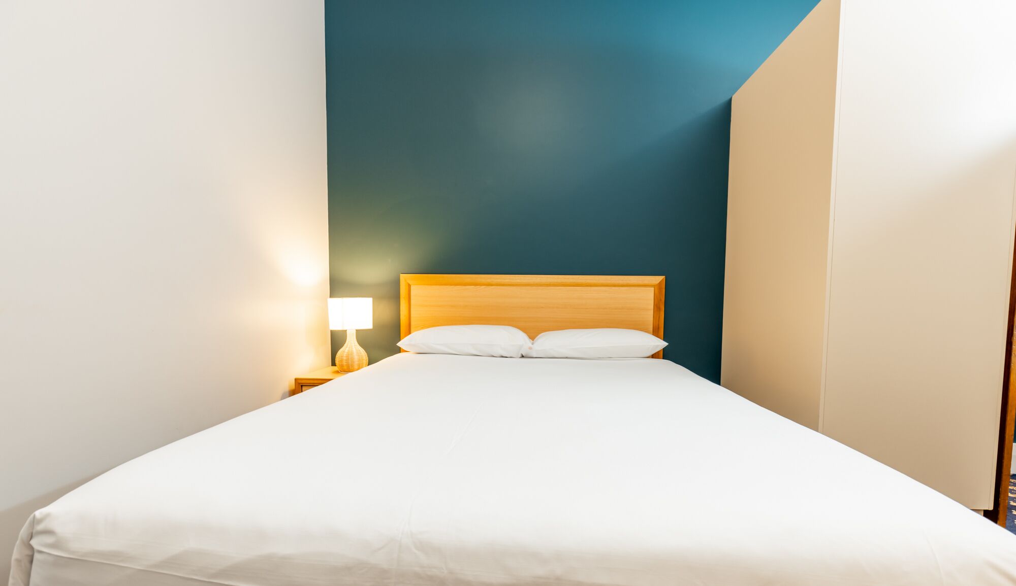 Een netjes opgemaakt bed met witte lakens in een kamer met een blauwe accentmuur en een houten hoofdbord.