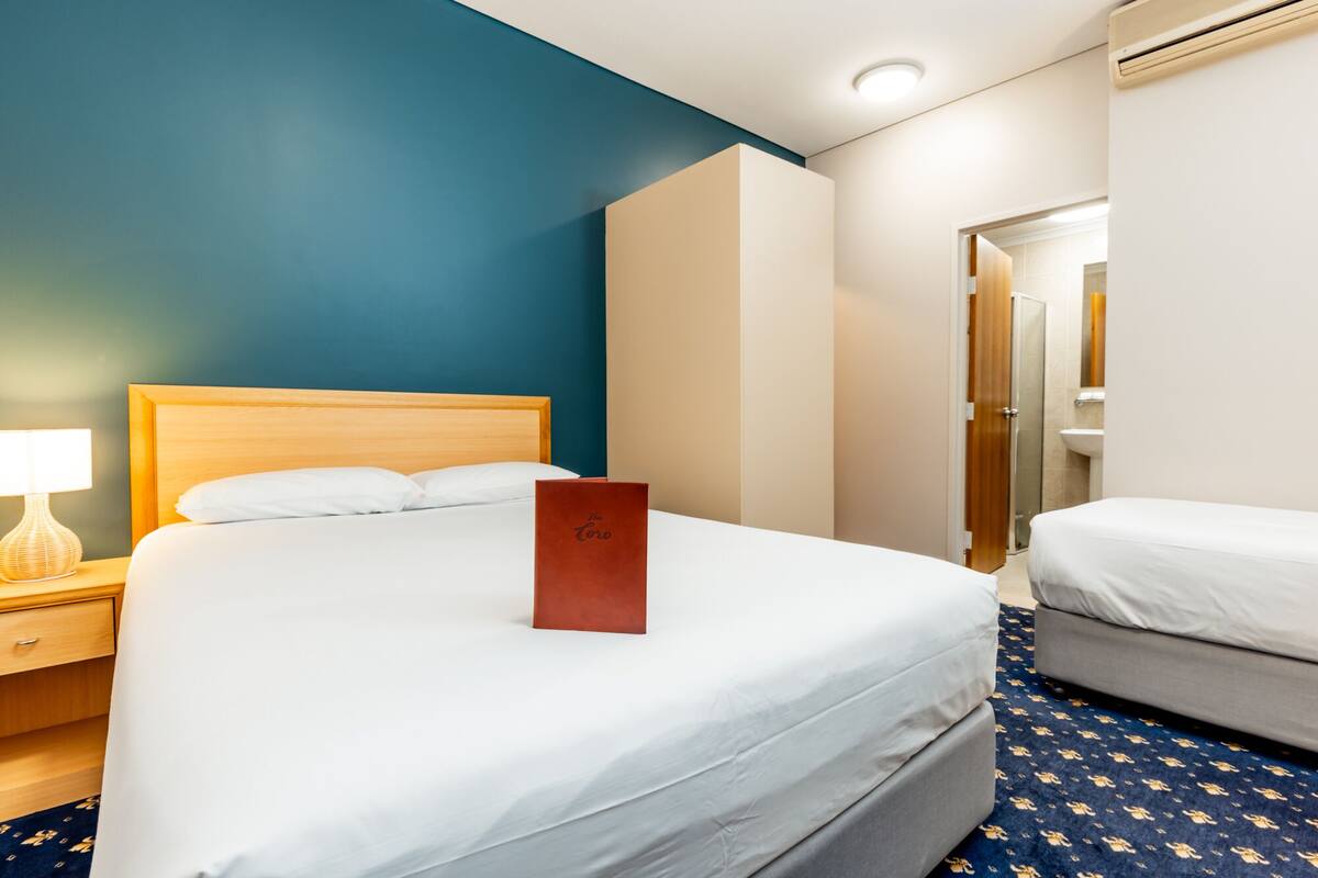 A hotel room with a large bed, a smaller bed, a nightstand with a lamp, and a bathroom visible through an open door.