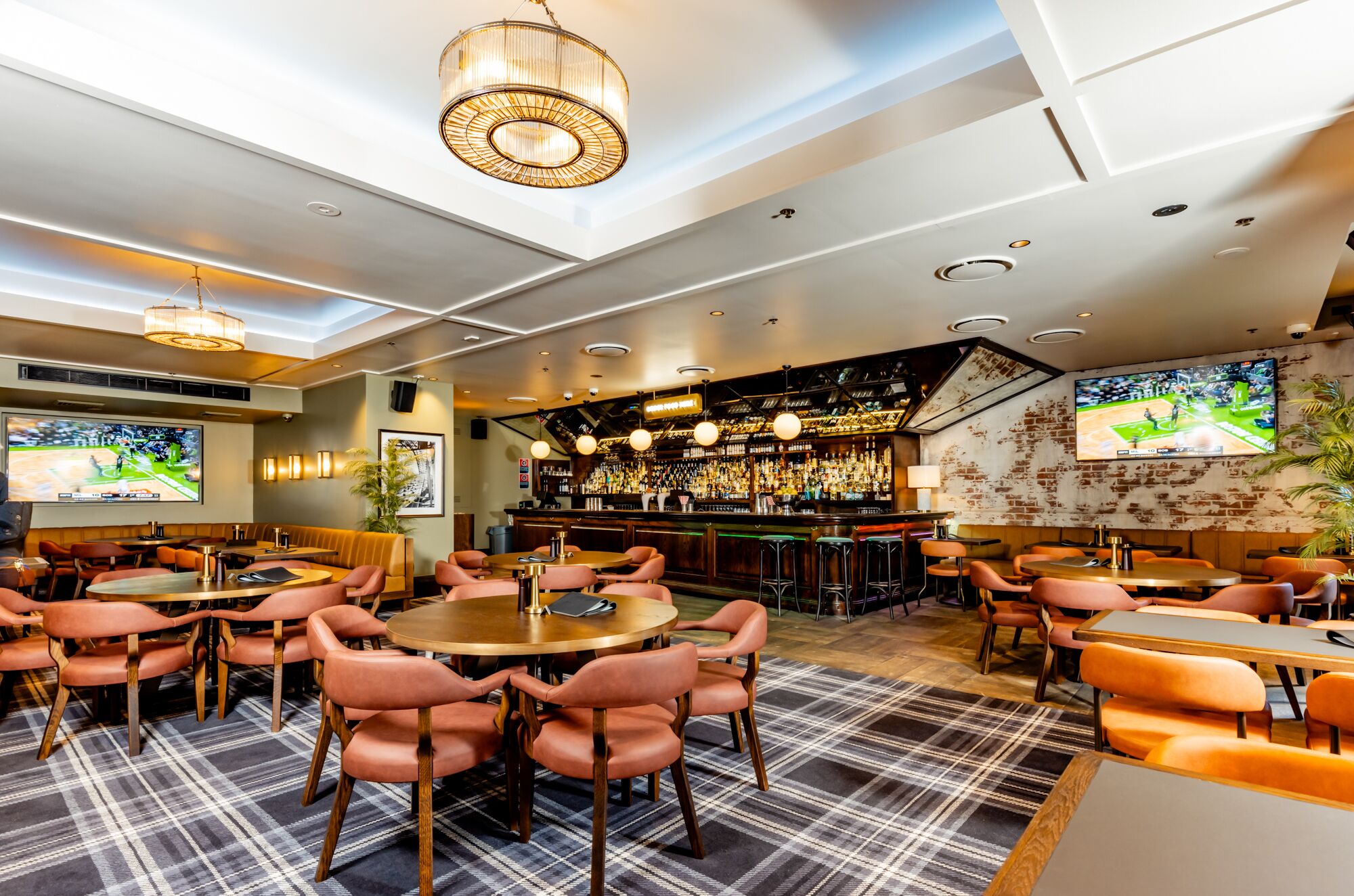 A bar area with multiple seating options, including round tables and bar stools, and a television screen displaying a sports event.