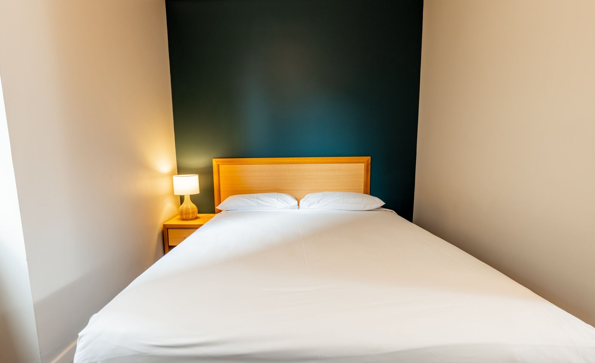 Een kleine hotelkamer met een enkel bed, een nachtkastje met lamp, en een donkere wand.