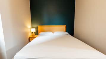 Een kleine hotelkamer met een enkel bed, een nachtkastje met lamp, en een donkere wand.