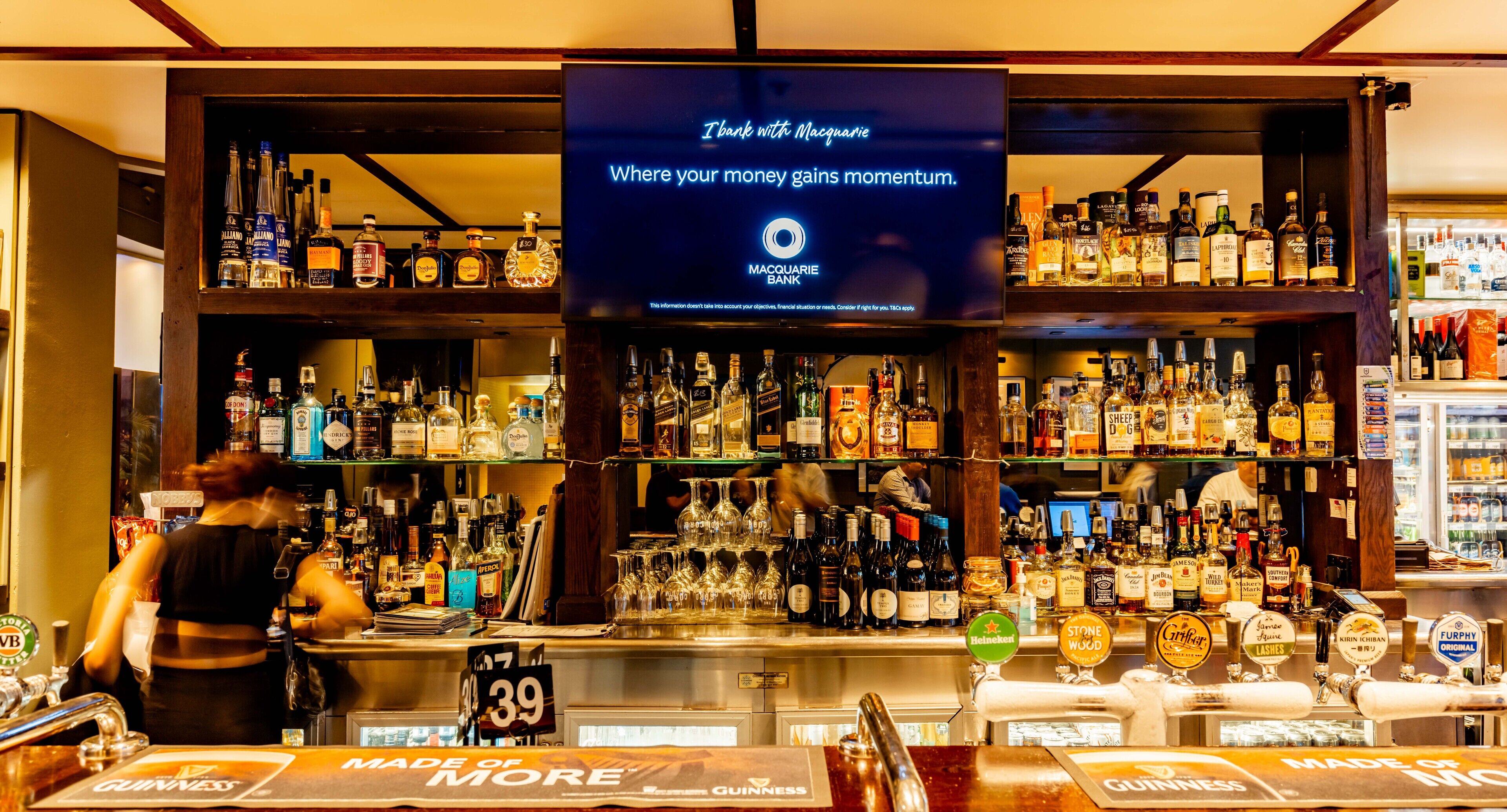 Een bar met een groot scherm waarop een promotiebericht staat, planken vol met verschillende drankflessen en meerdere biertaps.
