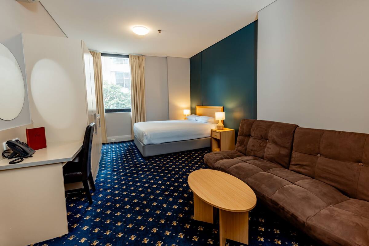 A hotel room with a bed, a sofa, a small table, a desk with a phone, and a window with curtains.
