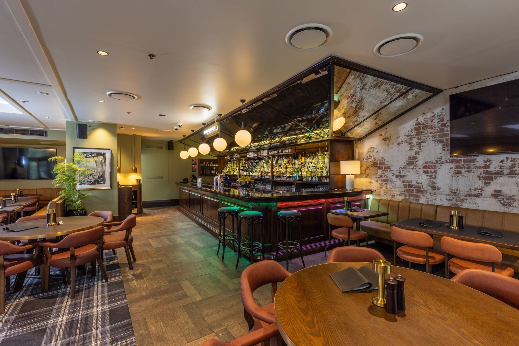 A bar area with a wooden counter, stools, and a variety of bottles. There are tables and chairs for seating, and a large screen on the wall.