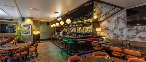 Una zona de bar con mostrador de madera, taburetes y una variedad de botellas. Hay mesas y sillas para sentarse, y una pantalla grande en la pared.