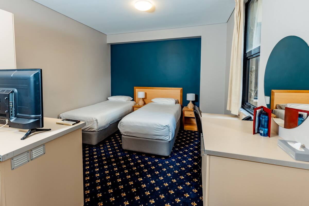 A hotel room with two single beds, a desk with a computer, a TV, and a window with curtains.